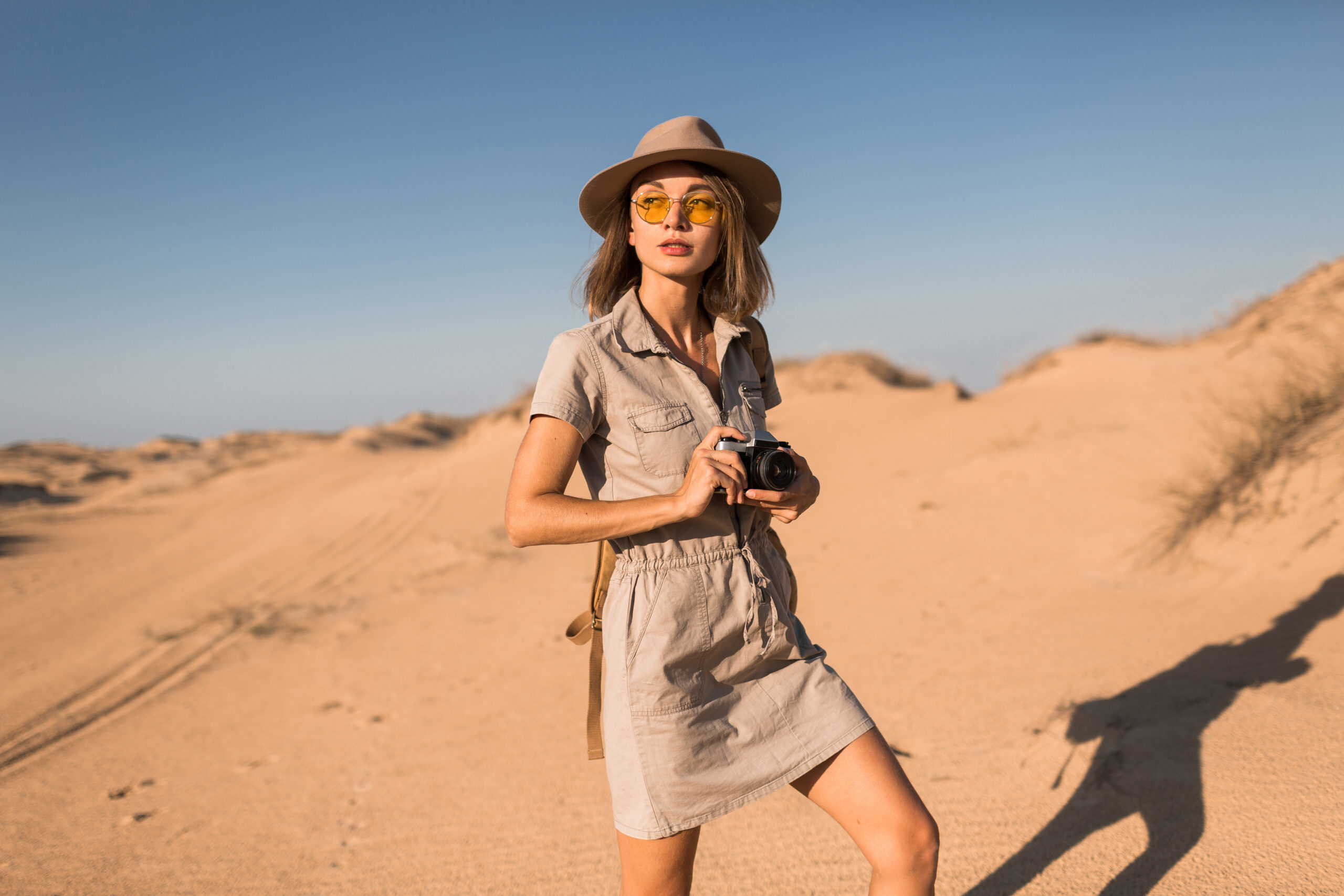 stylish young woman in khaki dress walking in desert, traveling in Africa on safari, wearing hat and backpack, taking photo on vintage camera, exploring nature, hot summer day, sunny weather